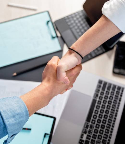 Two people shaking hands over laptops and documents in an office, symbolizing a business deal.
